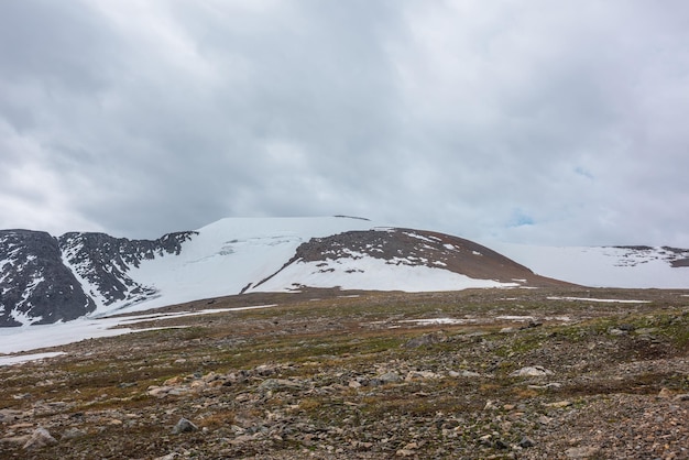 Dramatic landscape with stone field and large snowy mountain dome under gray cloudy sky Atmospheric scenery with high snow mountain in dome shape in center at cloudy weather Overcast in mountains