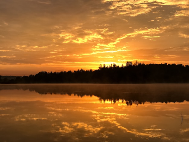 Dramatic orange sky at sunset over forest lake reflecting on still water surface, forest line silhouetted on horizon