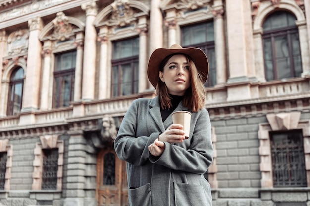 Dramatic portrait of a fashionable woman in an autumn coat and hat. Attractive girl holding cup of coffee on the background of urban architecture