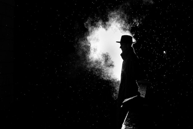 Drammatica silhouette di un uomo pericoloso in un cappello di notte sotto la pioggia in città nel vecchio stile crimine noir