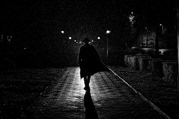 Dramatic silhouette of a man in a hat and raincoat walking\
through the city at night in the rain in retro noir style