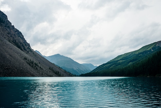 Dramatic view to vast mountain lake among giant mountains in rainy weather. Pines and larches on hillside near azure water. Overcast landscape with turquoise alpine lake. Atmospheric highland scenery.