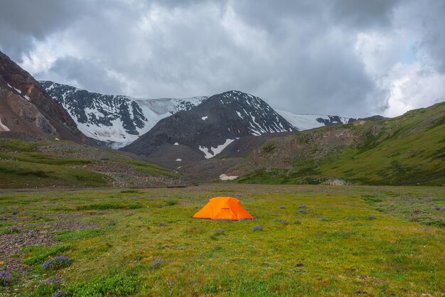 Dramatisch berglandschap met levendige oranje tent op gras in groene bergvallei met het oog op sneeuwbergen onder grijze bewolkte hemel Schilderachtig bewolkt landschap met sneeuwbergketen bij regenachtig weer