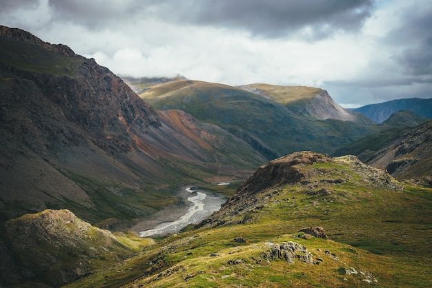 Dramatisch berglandschap met rivier in vallei tussen bonte rotsen in zonlicht onder bewolkte hemel. Toneel alpien groen landschap met bergrivier in diepe kloof onder bewolkte hemel bij veranderlijk weer