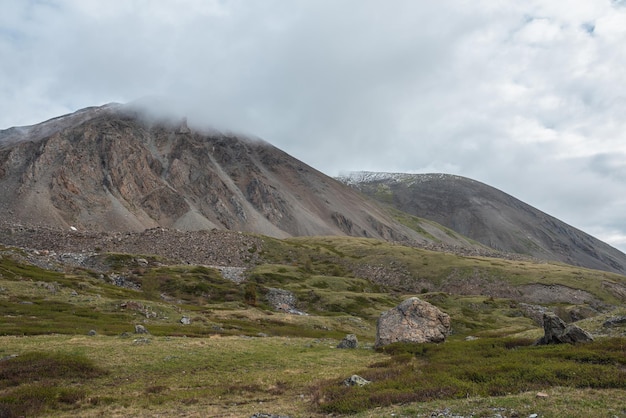 Dramatisch landschap met grote stenen op heuvels tegen hoge berg met scherpe rotsen in regenachtige lage wolken somber landschap met grote rotsachtige bergketen in lage grijze bewolkte lucht bij somber regenachtig weer