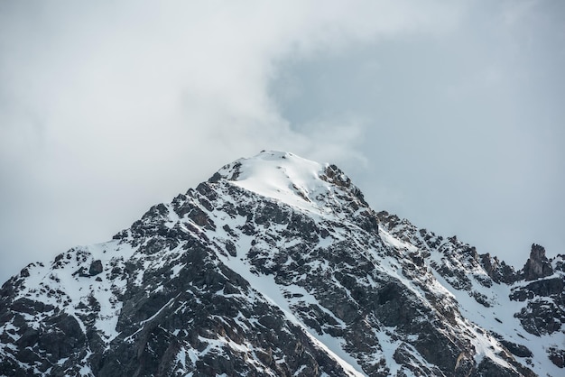 Dramatisch landschap met zonovergoten hoge besneeuwde bergtop in bewolkte lucht Witte sneeuw op zwarte rotsen in wolkenlucht Geweldige sneeuwbergtop in zonlicht bij wisselvallig weer Ingesneeuwde berg in wolken
