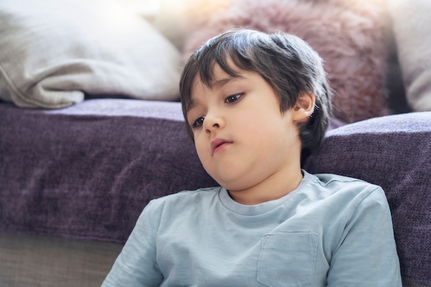 Dramatisch portret Eenzaam kind droevig gezicht zittend op de vloer, kind alleen zittend naast de bank in de woonkamer met diep in gedachten neergekeken, Jongen verveelt zich thuis tijdens zelfisolatie, Quarantaineconcept