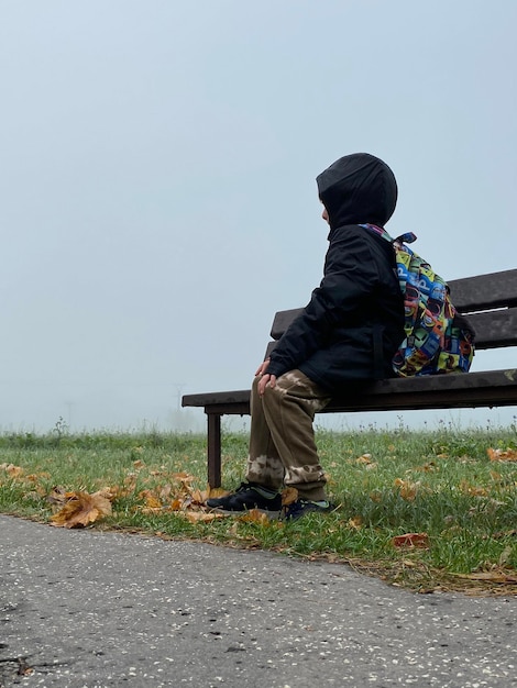 Dramatische scène van een kind met een rugzak alleen zittend op een bankje in een koude en mistige winterdag wachtend op zijn moeder of vader Jongen is verdwaald in het donker