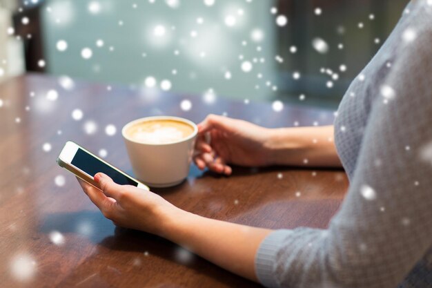 dranken, mensen, technologie en communicatie concept - jonge vrouw met smartphone koffie drinken in een café over sneeuw