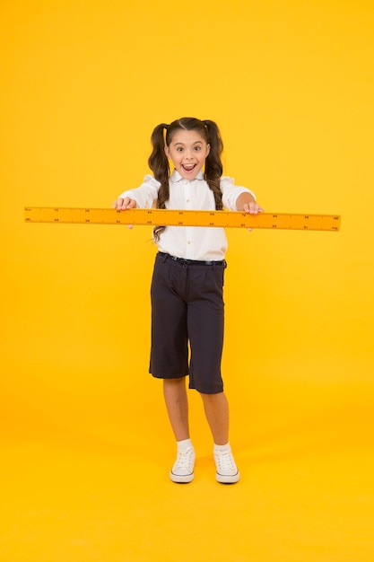 Drawing straight lines at geometry Happy small girl holding ruler for geometry lesson on yellow background Little child learning geometry Cute schoolchild preparing geometrical tool for geometry