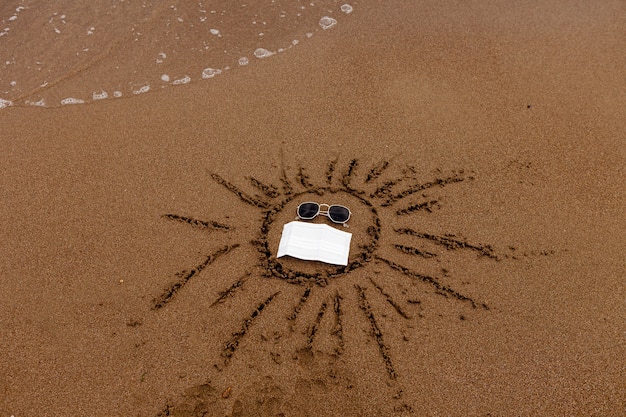 Disegnare occhiali da sole e una maschera medica sulla sabbia della spiaggia riposano in mare e sulla costa durante il ...