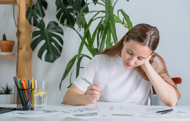 Drawings made in pencil are on the table a young woman of\
european appearance draws with a pencil on white paper graphics the\
process of drawing with a pencil at home at a white table