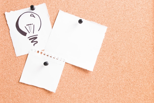 Drawn light bulb on a white sheet attached to a cork board with pin with other papers