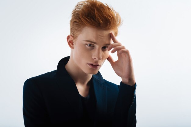 Dreamboat. Attractive serious red-headed young man wearing a black jacket and touching his forehead and thinking