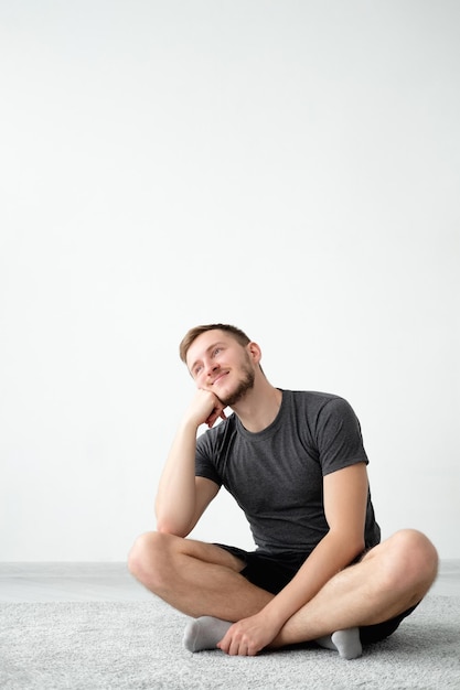 Dreamful man Happy selfisolation Plans goals Smiling guy in tshirt shorts sitting floor in lotus pose hands on cheek on light wall interior background