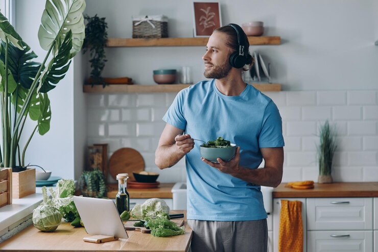 Photo dreamful young man in headphones eating fresh salad while leaning at the kitchen counter at home