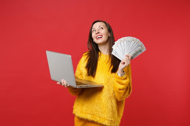 Dreamful young woman in fur sweater looking up hold laptop pc computer, fan of money in dollar banknotes, cash money isolated on red background. People emotions, lifestyle concept. Mock up copy space.