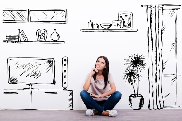 Dreaming about new apartment. Cheerful young woman smiling while sitting on the floor against white background with drawn home interior