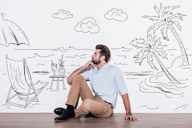 Dreaming about vacation. Young handsome man keeping hand on chin and looking away while sitting on the floor with illustration of resort in the background