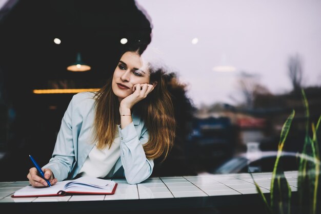 Dreaming beautiful hipster girl looking at window cafeteria while writing romantic essay