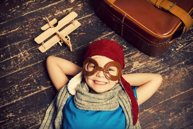 Dreaming of a big sky. Top view of happy little boy in pilot headwear and eyeglasses lying on the hardwood floor and smiling while wooden planer and briefcase laying near him