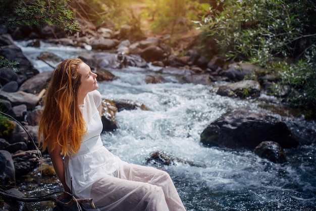 Sognare una donna romantica in abito bianco seduta sulla riva del fiume di montagna tra alberi verdi. giovane ragazza che alza il viso al sole sullo sfondo della natura. armonia concetto di stile di vita felice.