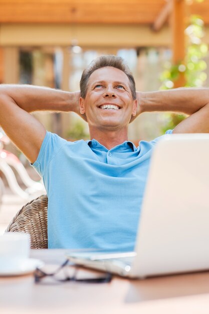 Dreaming of vacation. Relaxed mature man holding hands behind head and smiling while sitting at the table outdoors with laptop on it