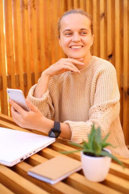 Dreaming woman wearing beige sweater sitting in outdoor cafe working on laptop resting using mobile phone looking away with cheerful expression