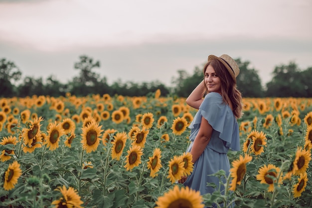 Dreaming young woman in blue dress and hat in a field of sunflowers at summer, view from her side. Looking forward. copy space