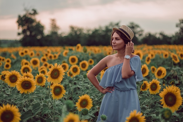 Foto sognando la giovane donna in abito blu e cappello che tiene i capelli con una mano in un campo di girasoli in estate, vista frontale. guardando di lato. copia spazio