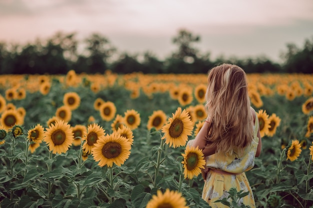 Dreaming young woman in yellow dress holding hairs with hands in a field of sunflowers at summer, view from her back. Looking to the side. copy space