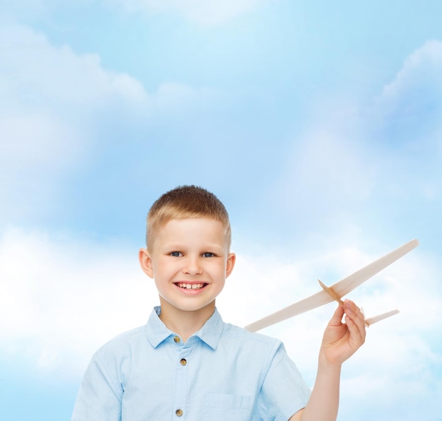 dreams, future, hobby and childhood concept - smiling little boy holding wooden airplane model in his hand over blue sky background