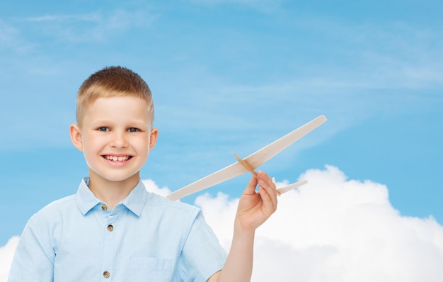 dreams, future, hobby and childhood concept - smiling little boy holding wooden airplane model in his hand over blue sky background