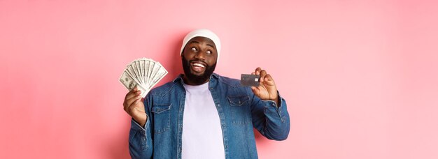 Dreamy africanamerican man showing credit card and dollars thinking about shopping and smiling stand