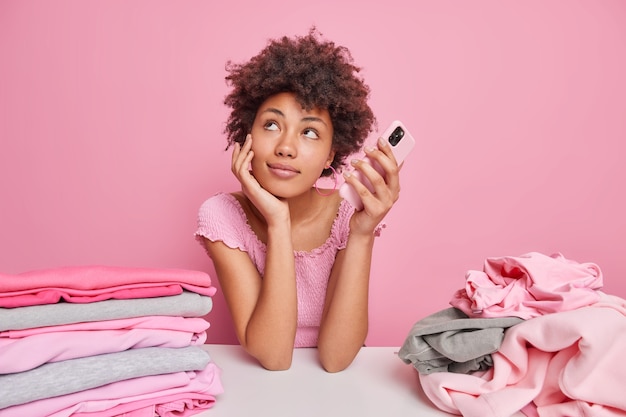 Dreamy Afro American woman folds clean clothes after laundry leans at white table holds mobile phone waits for call looks thoughtfully aside poses against pink wall stacks of clothing around