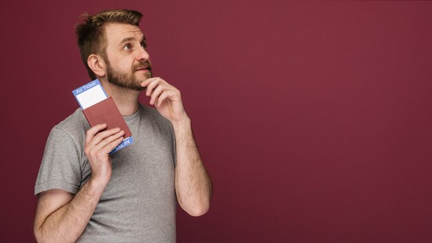 Dreamy bearded man in a t-shirt holding air tickets and passport against a ruby background