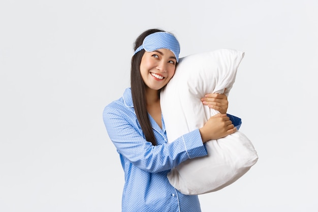 Dreamy beautiful asian girl in blue pajamas and sleeping mask hugging pillow and smiling pleased as looking upper left corner imaging something in bed, picturing, using imagination.