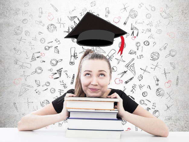 Foto dreamy blonde vrouw zit aan een tafel met haar hoofd op een stapel boeken. betonnen muur achtergrond met onderwijs iconen en een afstuderen hoed boven haar.
