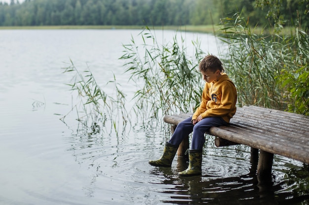 Dreamy boy sitting on the wooden pier at pond and play with water