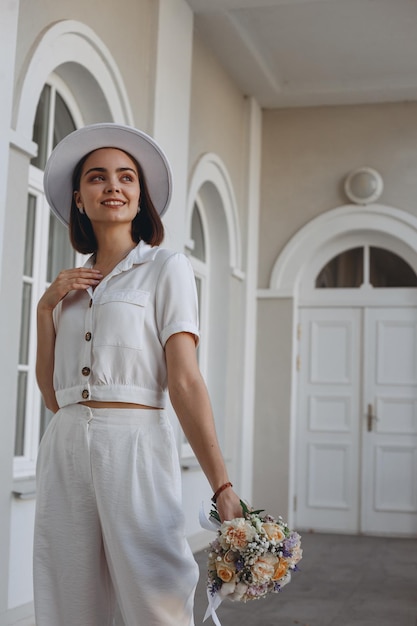 Dreamy bride in white fancy hat and trousers and with wedding flower bouquet