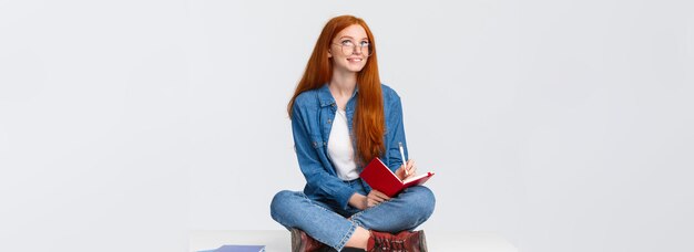 Dreamy and creative cute diligent female student with red hair in glasses sitting on floor with legs