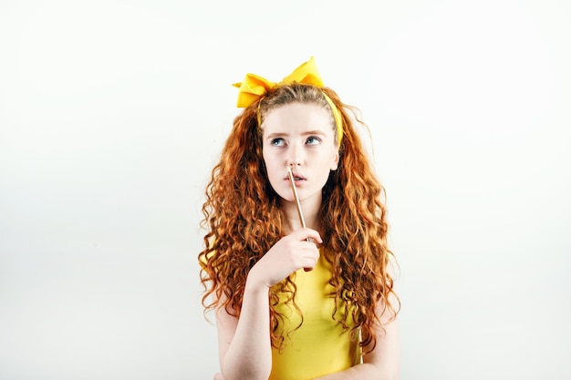 Dreamy curly ginger girl with a yellow bow on her head holding pencil near her mouth and thinking while standing on the white background