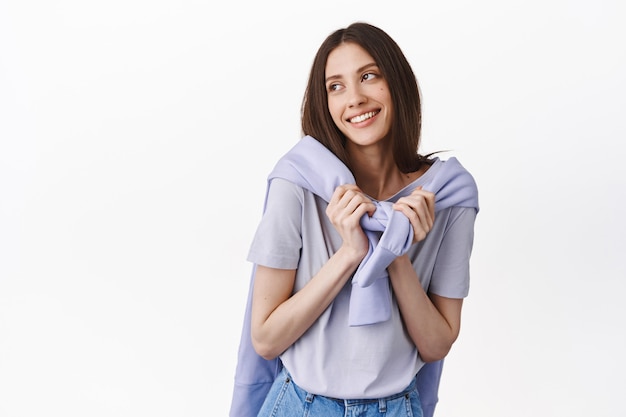 Dreamy and cute girl wrap sweatshirt around neck, looking aside at upper left corner with daydreaming face, romantic gaze, smiling and standing over white wall