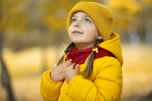 Dreamy cute little girl in stylish warm autumn clothes, posing outdoors in the golden city park and looking up. Pretty child girl in yellow outfit enjoying autumn season in the nature.