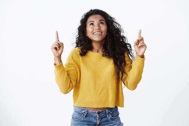 Sognante donna afroamericana sorridente carina in maglione giallo ansimando divertita, guarda e punta le dita verso l'alto stupita, vedi una fantastica opportunità, ride spensierata, muro bianco