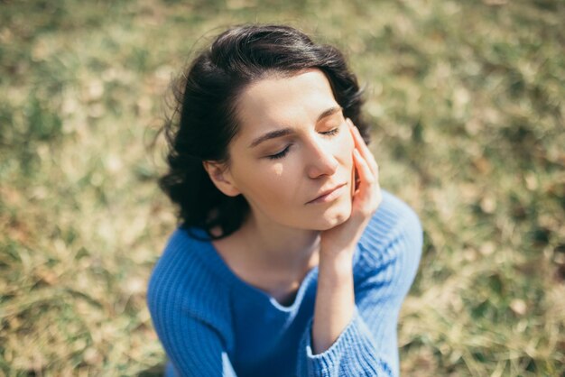 写真 顔に手を閉じて目を閉じた夢のような女性春や夏に公園に座って外で夢を見ている風の強い髪を持つ魅力的なブルネットの白人の若い女性の肖像画ライフスタイルのコンセプト