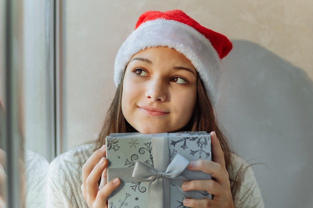 dreamy girl in a Christmas hat with a gift box by the window closeup