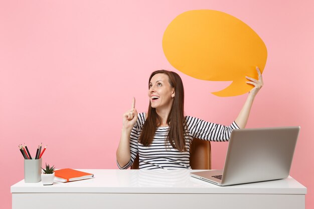 Dreamy girl holding yellow empty blank Say cloud speech bubble work at white desk with pc laptop isolated on pastel pink background. Achievement business career concept. Copy space for advertisement.