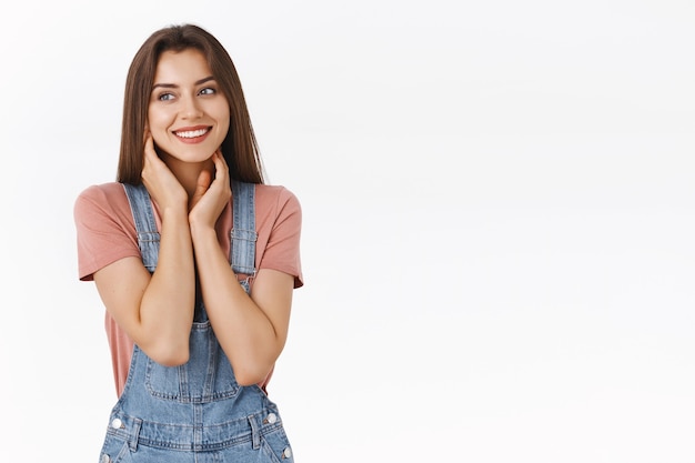 オーバーオール、Tシャツ、両手で首に優しく触れ、白い背景の上に控えめまたは恥ずかしがり屋、いちゃつく、立っているコケティッシュを見て微笑んでいる夢のような、素敵でフェミニンな美しい女性