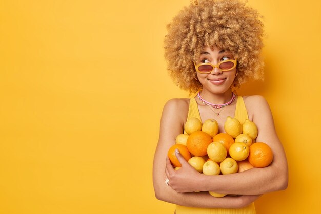 Dreamy lovely woman looks pensively away smiles gently embraces\
big heap of fresh oranges and lemons wears sunglasses enjoys summer\
time isolated over yellow background empty space for your text